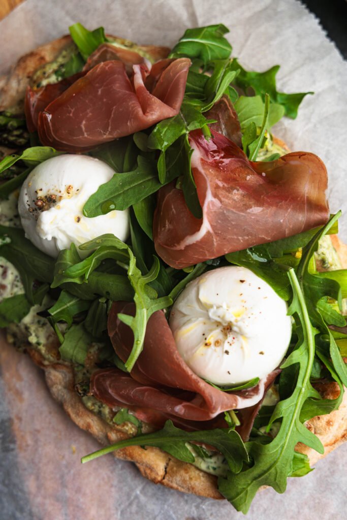 Pinsa with wild garlic crème fraîche spread, green asparagus, topped with arugula, wild garlic leaves, slices of dry-cured ham, and two small burratas. Served on baking paper over a wooden board, styled with a basket of fresh wild garlic and seasoning bowls. A vibrant spring dish perfect as a light dinner or appetizer.