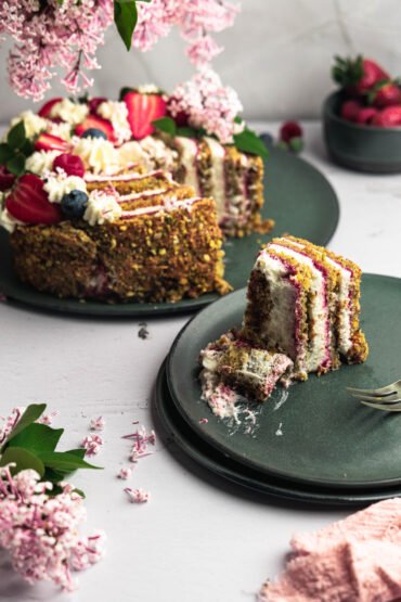 Pistachio cake with raspberry filling and cream, decorated with fresh strawberries, blueberries, and pink lilac flowers. One slice is served on a plate, surrounded by scattered blossoms and a pink cloth.