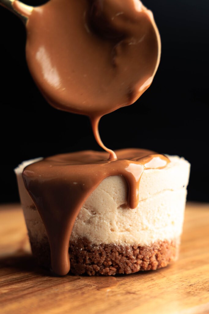 Close-up of creamy Lotus Biscoff spread being poured over a mini Lotus cheesecake with a biscuit base on a wooden board.