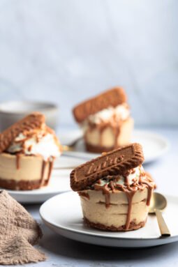 Mini Lotus cheesecaky s vyšľahanou smotanou, polevou z Lotus Biscoff krému a sušienkou Lotus, podávané na bielych tanieroch.