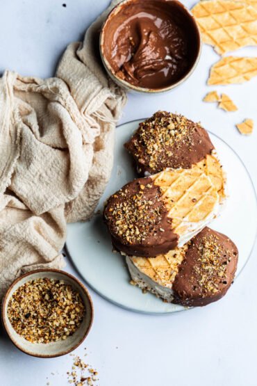 In Schokolade getauchte und mit gerösteten Nüssen bestreute Eiscreme-Sandwiches, serviert auf einem weißen Teller.