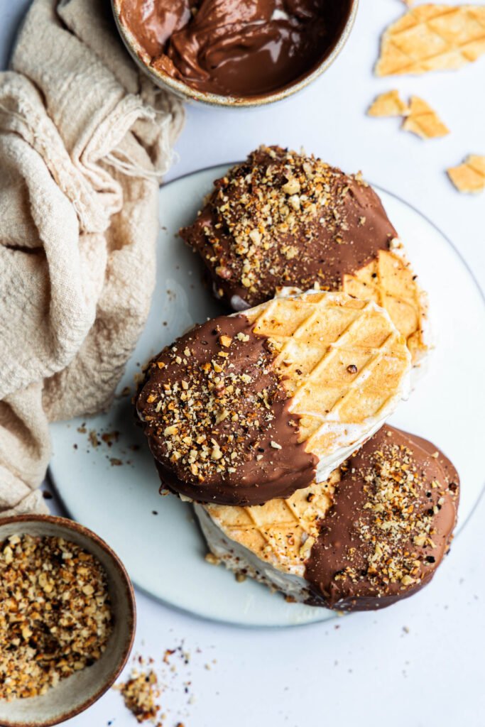 Top view of ice cream sandwiches coated in chocolate and decorated with chopped roasted nuts.