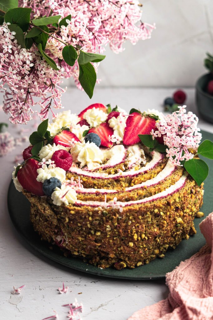 „Zarter Pistazienkuchen mit Himbeerschicht und luftiger Creme – dekoriert mit frischen Erdbeeren, Heidelbeeren und blühendem Flieder. Perfekt für Frühlingsfeste!“
