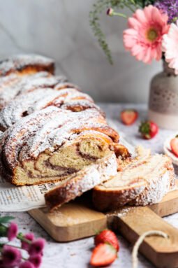 Nakrájaná kokosovo-jahodová babka posypaná práškovým cukrom, servírovaná na drevenej doske s kvetmi a jahodami.