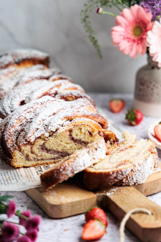 Nakrájaná kokosovo-jahodová babka posypaná práškovým cukrom, servírovaná na drevenej doske s kvetmi a jahodami.