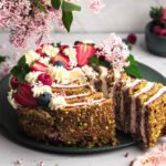 Jemný pistáciový koláč s malinovou vrstvou a nadýchaným krémom – ozdobený čerstvými jahodami, čučoriedkami a kvitnúcim orgovánom. Perfektný na jarné oslavy!