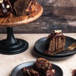 Marbled coffee bundt cake with a glossy chocolate glaze and chopped hazelnuts, served on a wooden cake stand with individual slices plated on black dishes.