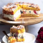 vrstvená torta plnená šľahačkou a lesným ovocím, podávaná na drevenom stojane s pozlátenou vidličkou a pivonkami v pozadí.