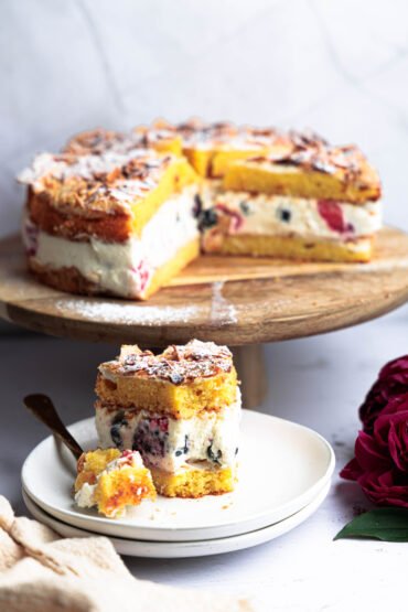 vrstvená torta plnená šľahačkou a lesným ovocím, podávaná na drevenom stojane s pozlátenou vidličkou a pivonkami v pozadí.