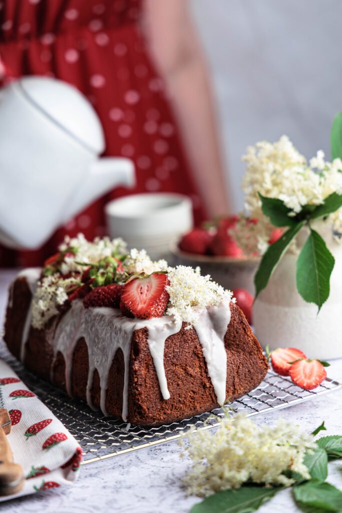 Elderflower-glazed loaf cake topped with strawberries and fresh elderflowers on a cooling rack, a teapot and summer table setting in the background.