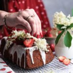 Eine Hand dekoriert den Holunder-Zitronen-Kuchen mit frischen Holunderblüten und Erdbeeren, im Hintergrund ein rotes Kleid.