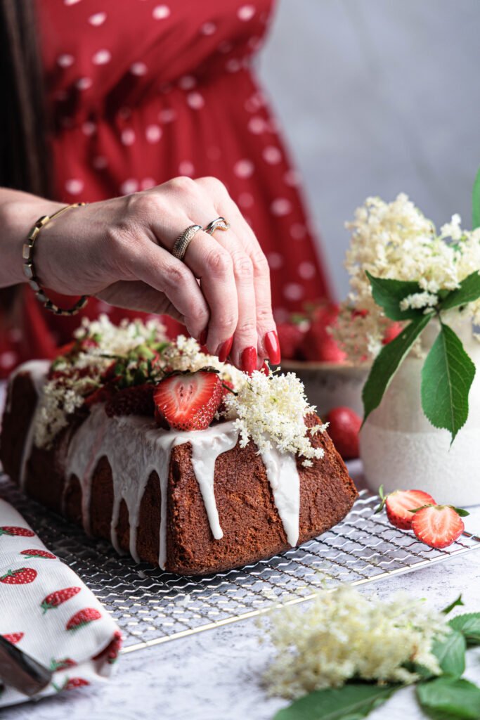 Eine Hand dekoriert den Holunder-Zitronen-Kuchen mit frischen Holunderblüten und Erdbeeren, im Hintergrund ein rotes Kleid.