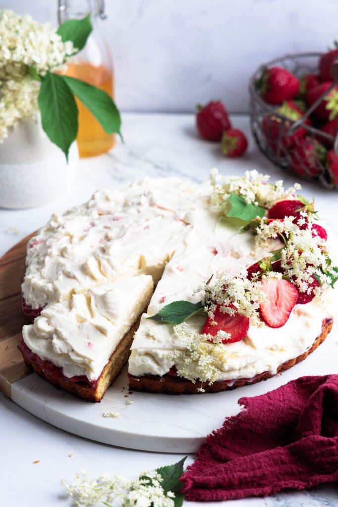 Elderflower strawberry tart with whipped mascarpone cream, decorated with fresh strawberries and elderflowers on a wooden cake board.
