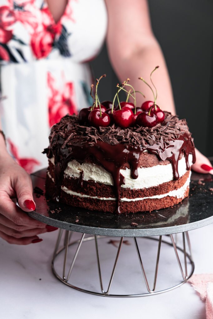 A woman in a floral dress holding a chocolate cherry cake topped with chocolate ganache and fresh cherries.
