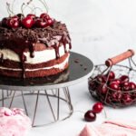 Čerešňová torta s čokoládovými hoblinami a polevou na kovovom stojane, obklopená čerešňami, kvetmi a ružovou utierkou.