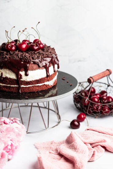Čerešňová torta s čokoládovými hoblinami a polevou na kovovom stojane, obklopená čerešňami, kvetmi a ružovou utierkou.