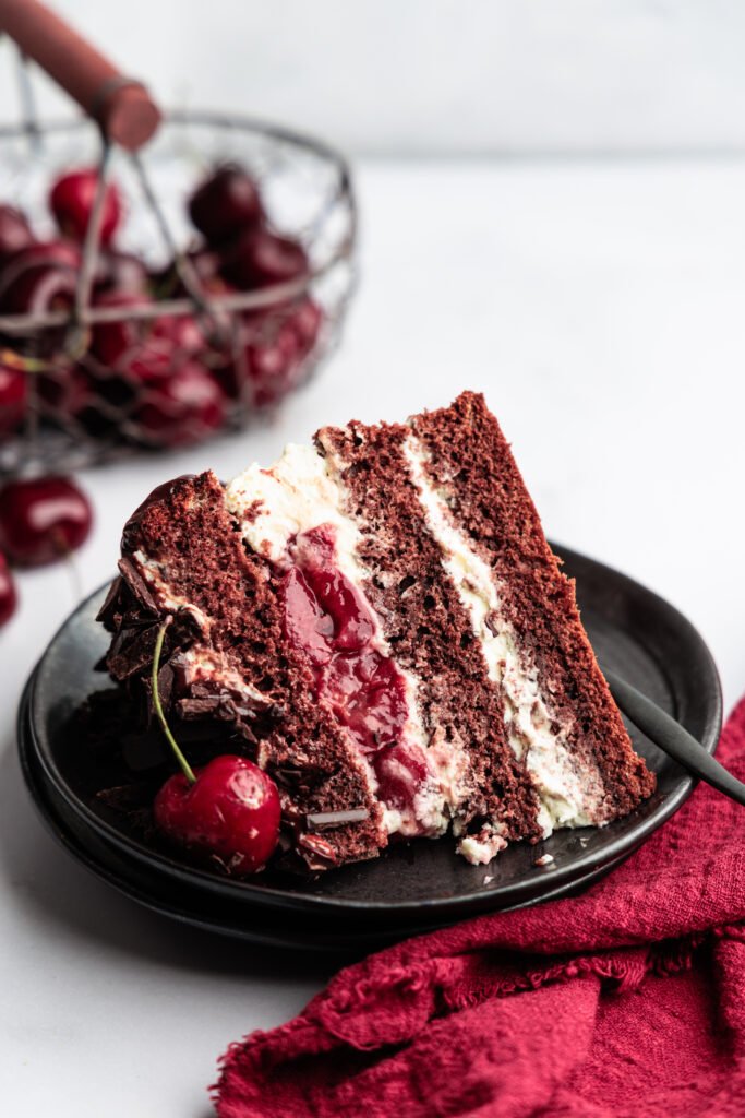 Ein Stück Schoko-Kirsch-Torte mit Sahne und frischen Kirschen auf einem schwarzen Teller.
