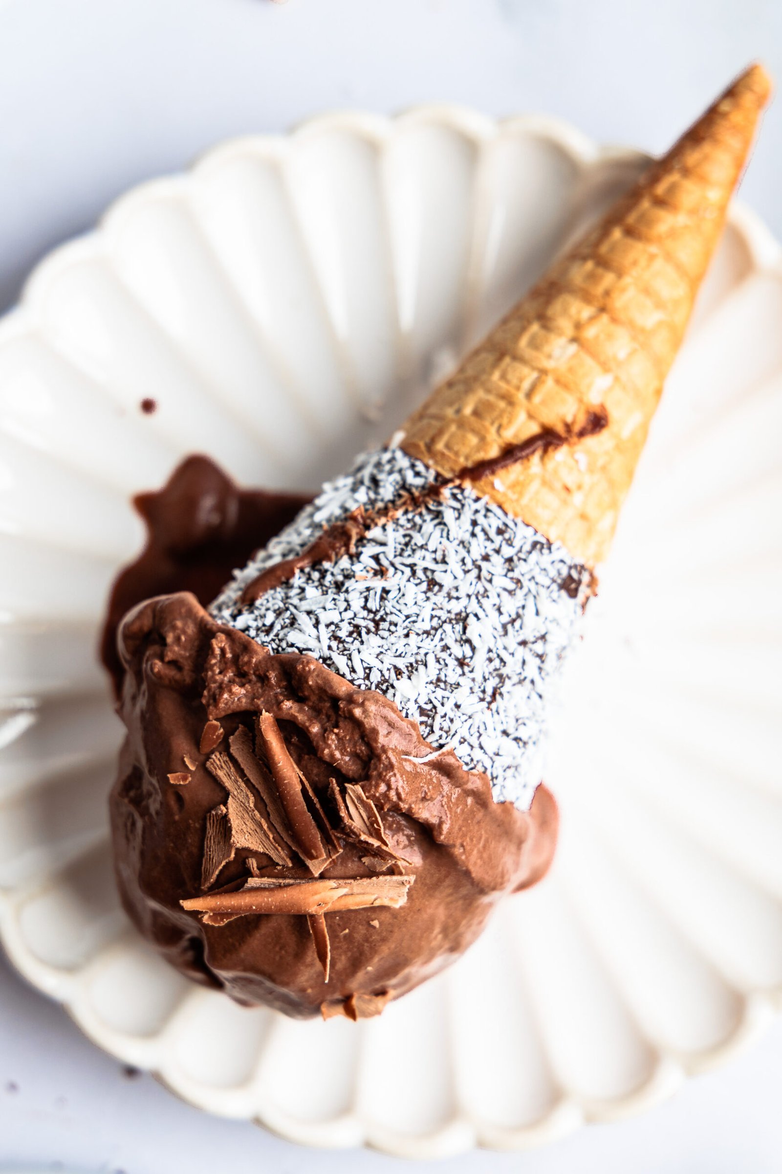 Close-up of chocolate ice cream in a coconut-covered cone, topped with chocolate flakes, served on a white plate.
